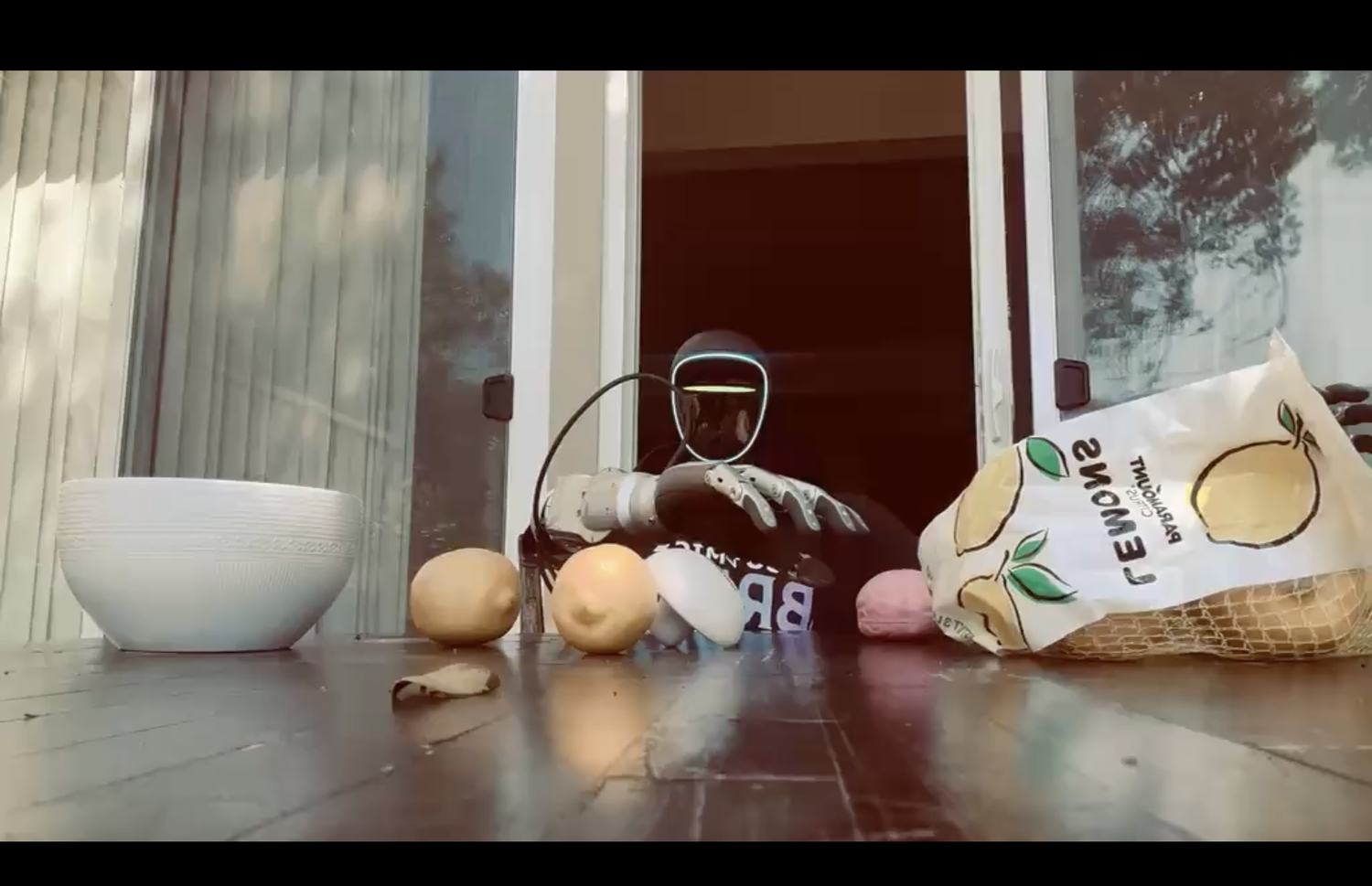 A humanoid robot in a Cosmic Brain shirt reaching toward produce scattered on a wooden deck beside a white bowl.